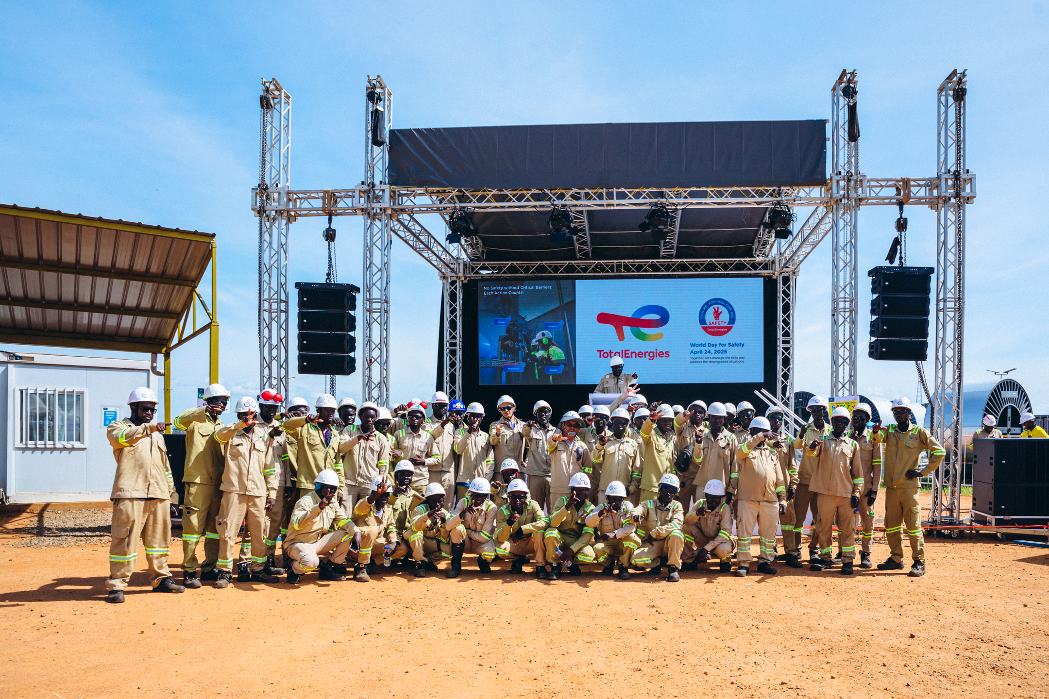 Group of workers in beige uniforms and white helmets posing outdoors in front of a stage with a TotalEnergies World Day for Safety 2025 banner.