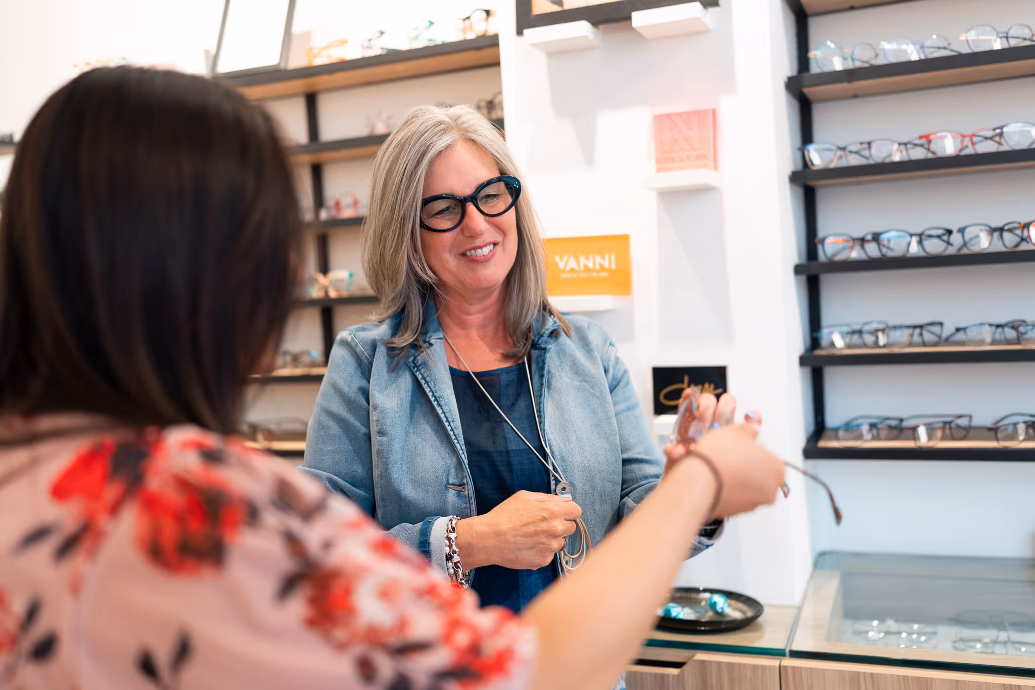 Optician helping a patient pick the perfect pair of glasses for their unique style; wall of glasses behind them