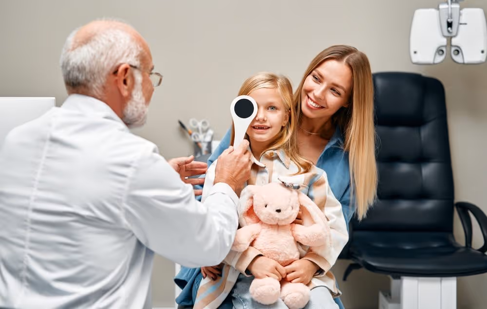 How Can Wellington Vision Care Improve Your Vision? Ophthalmologist closing one eye of a cute little girl while sitting on her mother's lap during a consultation. A child points to an eye chart during an eye test.