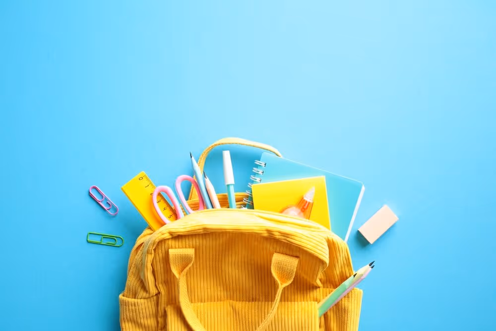 See the Good: Help Us Fill Backpacks for the Boys and Girls Club This Summer!  Back to School Concept. Brightly colored yellow backpack overflowing with essential school supplies such as notebooks, pens, pencils, scissors, and a ruler on a vibrant blue background. Flat lay.