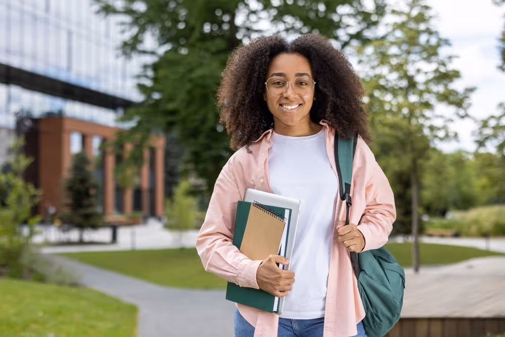 Wellington Eye Doctor: Caring for Your Eye Health. Young smiling woman student standing outside on campus. She holds notebooks and wears glasses, casual clothing, and a backpack. Beautiful day with green trees and modern buildings in background.