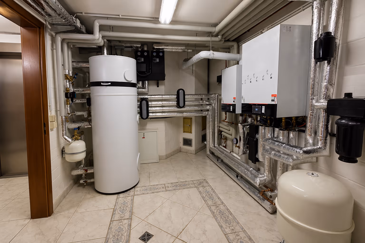 Technikraum mit neuer Heizungsanlage, isolierten Rohrleitungen und Tank in einem hell gefliesten Raum.