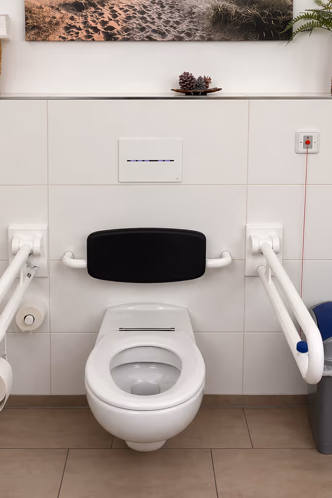 Behindertengerechte Toilette mit weißen Haltegriffen, schwarzer Rückenlehne, Wand-Tastaturspüler und Notrufknopf mit rotem Band.