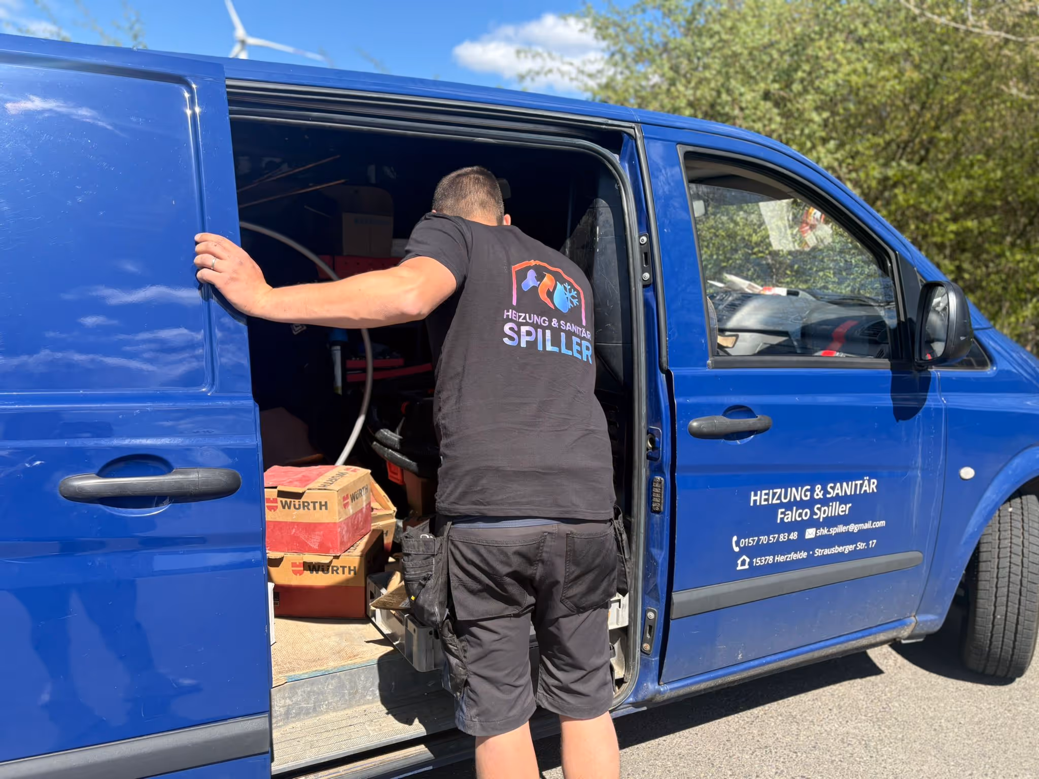 Handwerker in schwarzem T-Shirt mit der Aufschrift 'Heizung & Sanitär Spiller' läd Werkzeuge aus einem blauen Transporter.