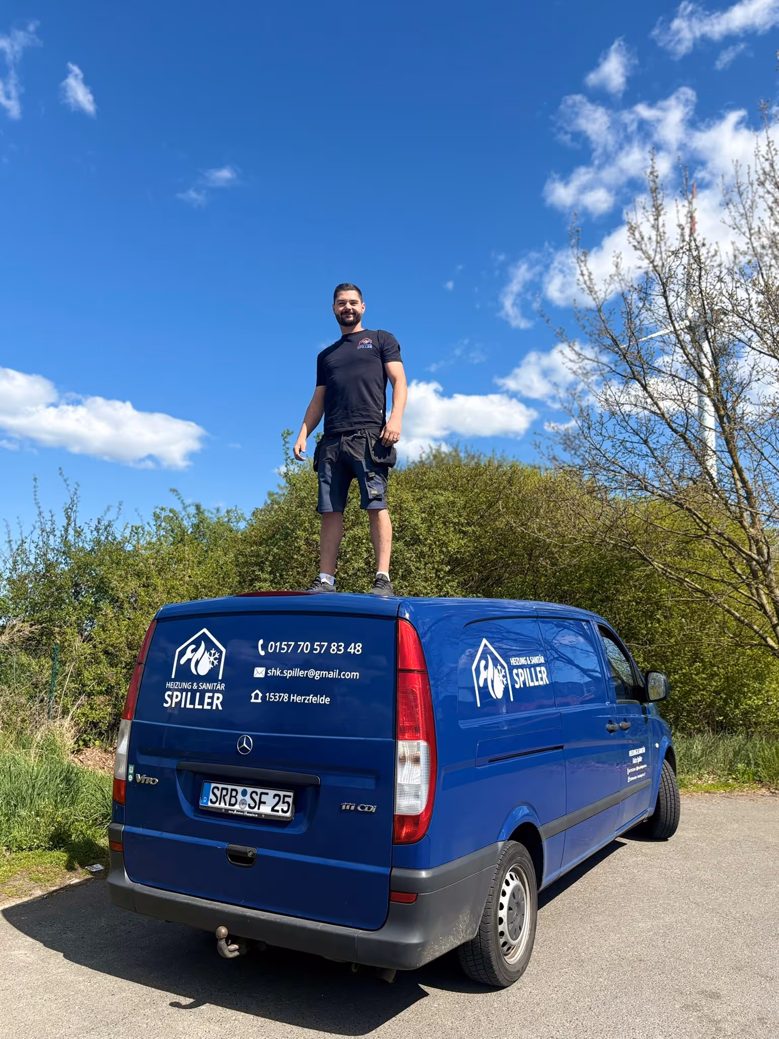 Mann in schwarzem T-Shirt und Shorts steht auf einem blauen Transporter mit Heizung und Sanitär Spiller Logo und Kontaktdaten, unter blauem Himmel mit einigen Wolken.