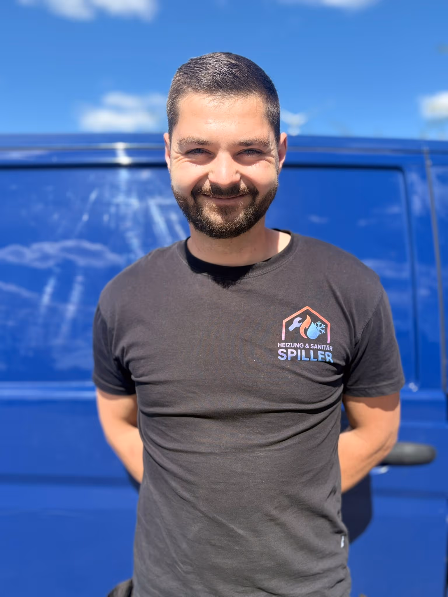Mann mit kurzem dunkelbraunem Haar und Bart lächelt vor einem blauen Fahrzeug, trägt schwarzes T-Shirt mit Logo ‚Heizung & Sanitär Spiller‘.