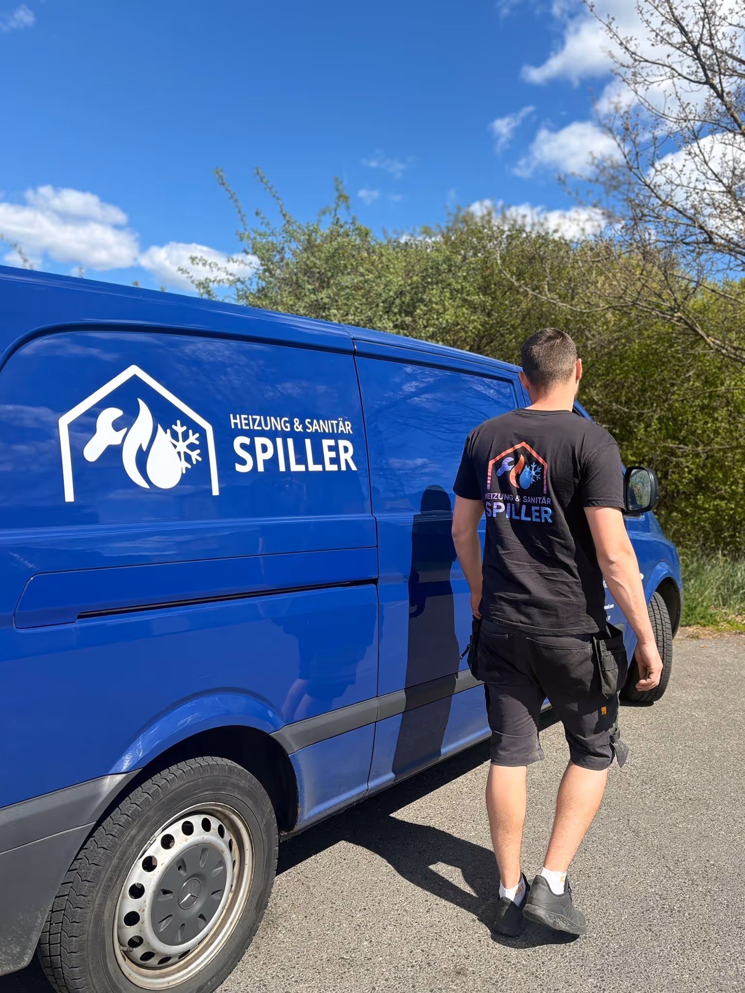 Ein Mann in schwarzem T-Shirt mit dem Logo von Heizung & Sanitär Spiller geht an einem blauen Transporter mit demselben Logo vorbei.