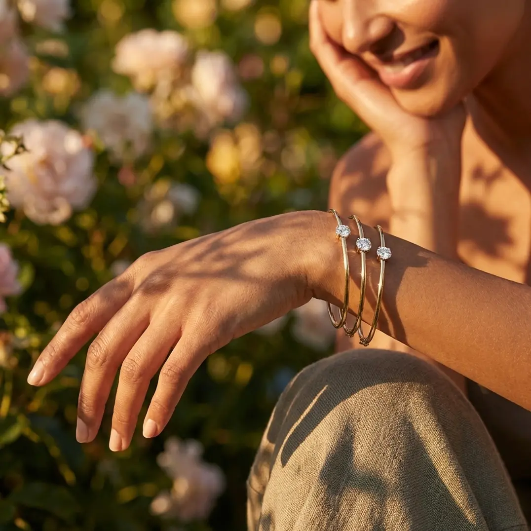 Close-up of a smiling woman wearing three gold bracelets with round diamond accents on her wrist, with a blurred floral background.