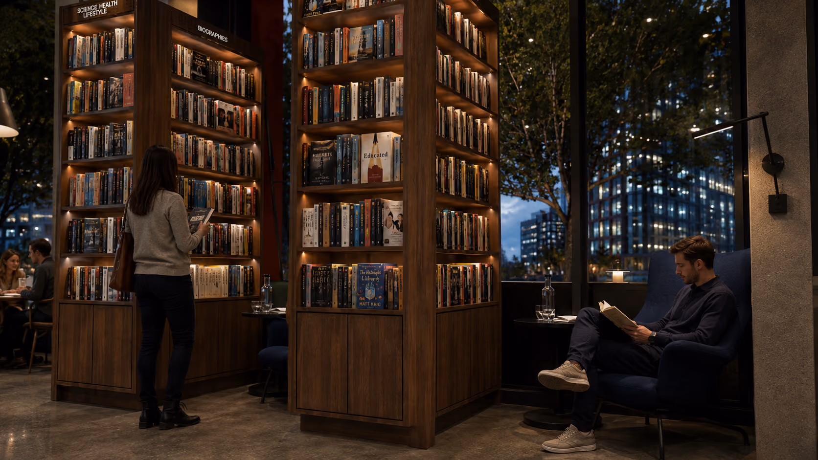 Woman browsing books in a warmly lit bookstore section labeled 'Science Health Lifestyle' and 'Biographies' while a man reads a book seated in a blue armchair near a large window with a cityscape view at night.