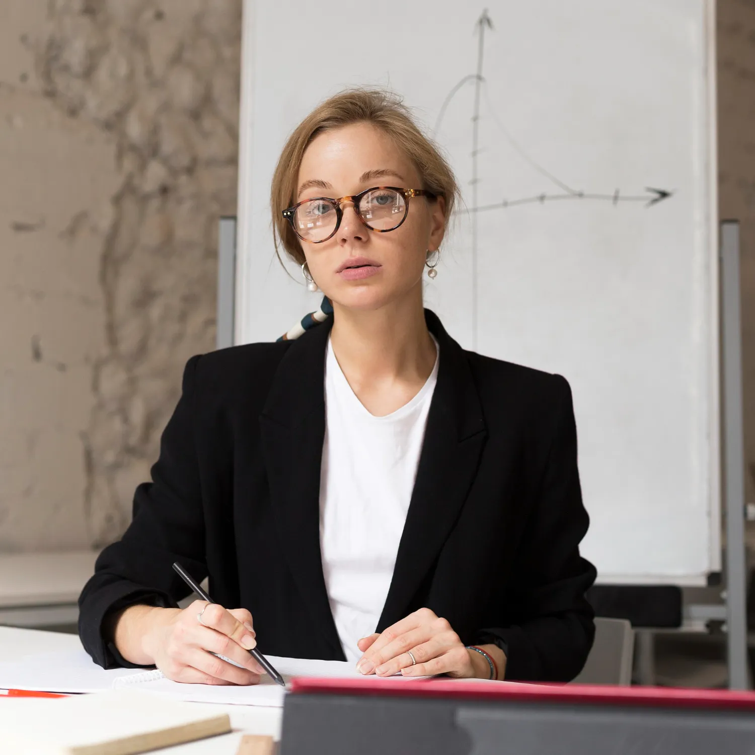 Woman wearing glasses and a black blazer writing on paper with a pen in front of a whiteboard with a graph.