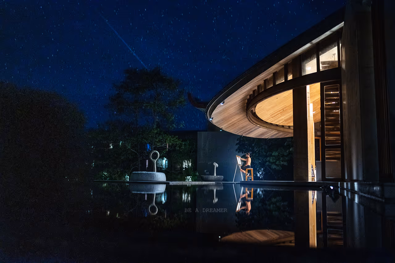 אדם יושב על שרפרף ומצייר על קנבס באור אור חם מחוץ למבנה עץ מודרני בלילה עם כיפת שמיים מכוכבים והשתקפות במים.