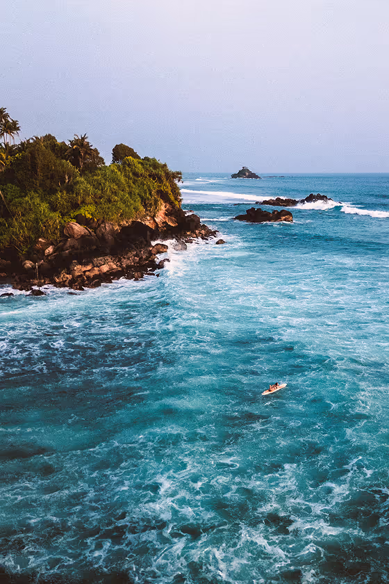 איש גולש על גלשן בגלים כחולים ליד חוף עם סלעים וצמחייה ירוקה.