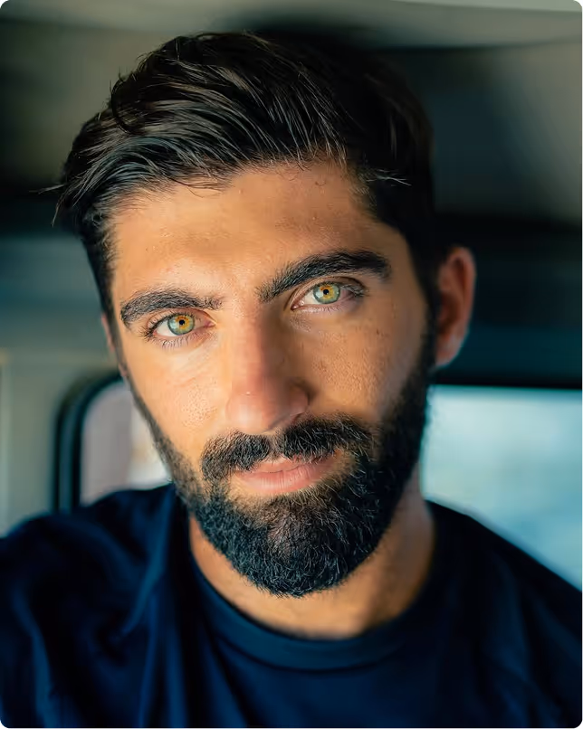 זכר צעיר עם שיער כהה, זקן ועיניים ירוקות מסתכל ישר למצלמה ברקע מטושטש.