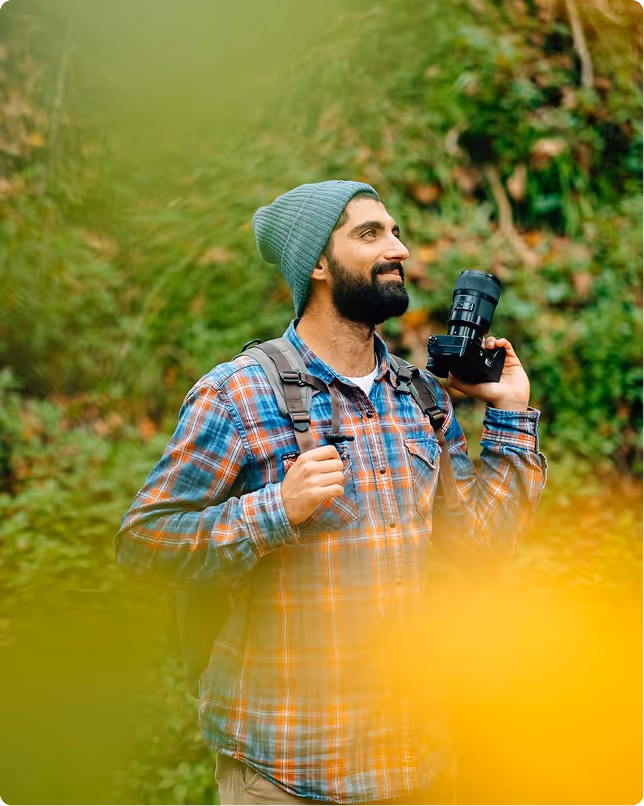 גבר עם כובע גרב וזקן מחזיק מצלמה ותרמיל בין צמחייה ירוקה וצהובה בטבע.