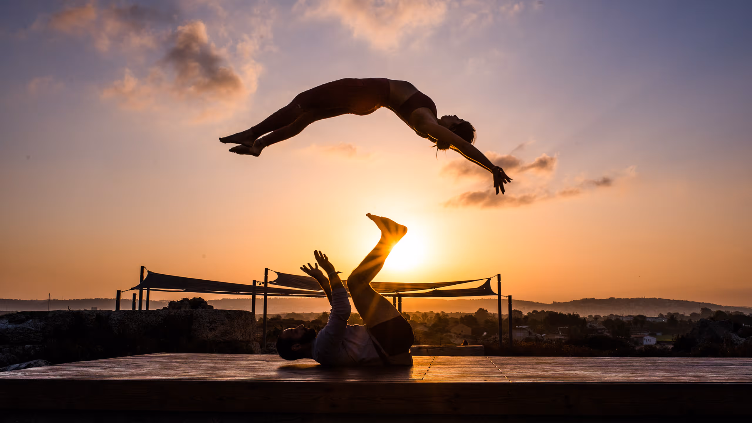 זוג מבצע תרגיל אקרובטיקה חיצונית בשקיעה עם האדם התחתון שוכב על הרצפה ומרימה את האדם העליון באוויר.