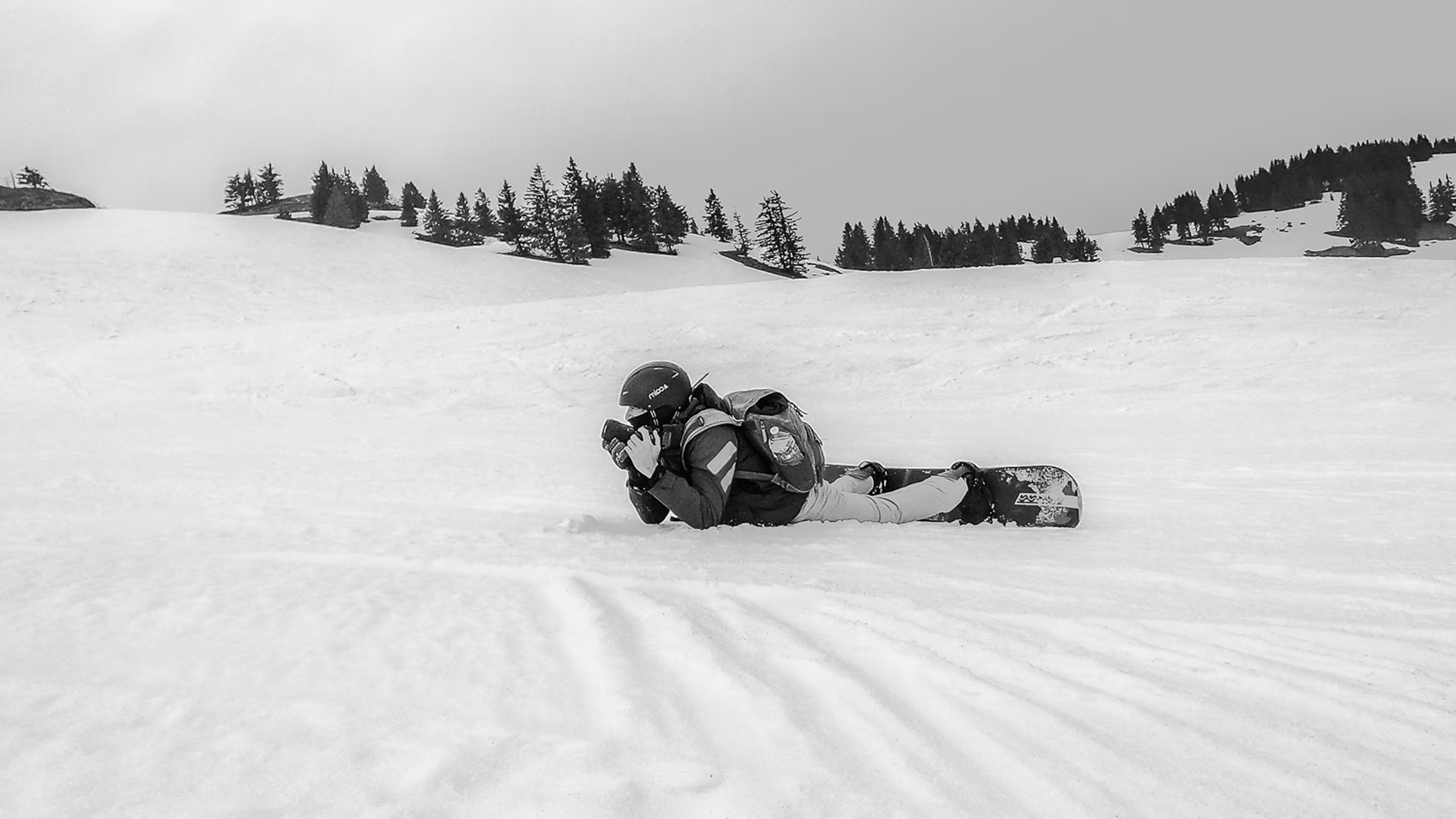 שלגייה שוכב על השלג עם סנובורד ומצלמה בידי, מוקף עצי מחט ברקע.