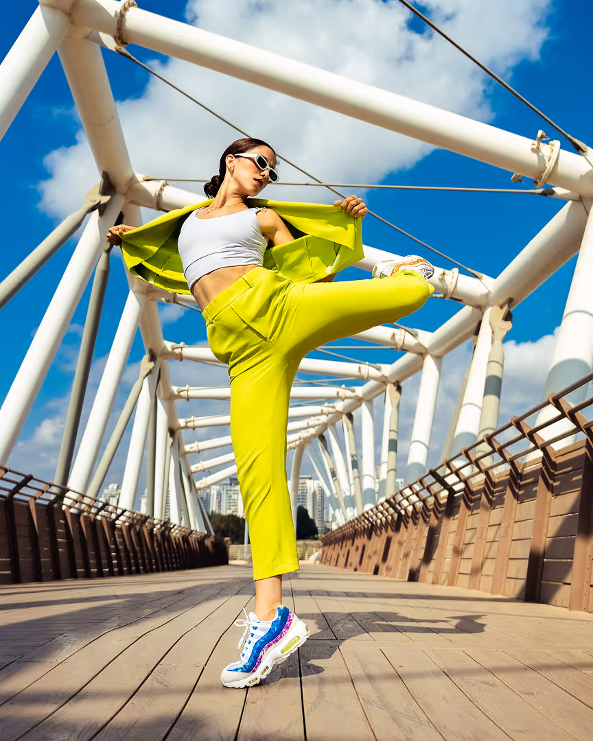 אישה בלבוש צהוב זוהר וצעיף לבן מבצעת בעיטה גבוהה על גשר פיקניק מעוצב תחת שמיים כחולים עם עננים.