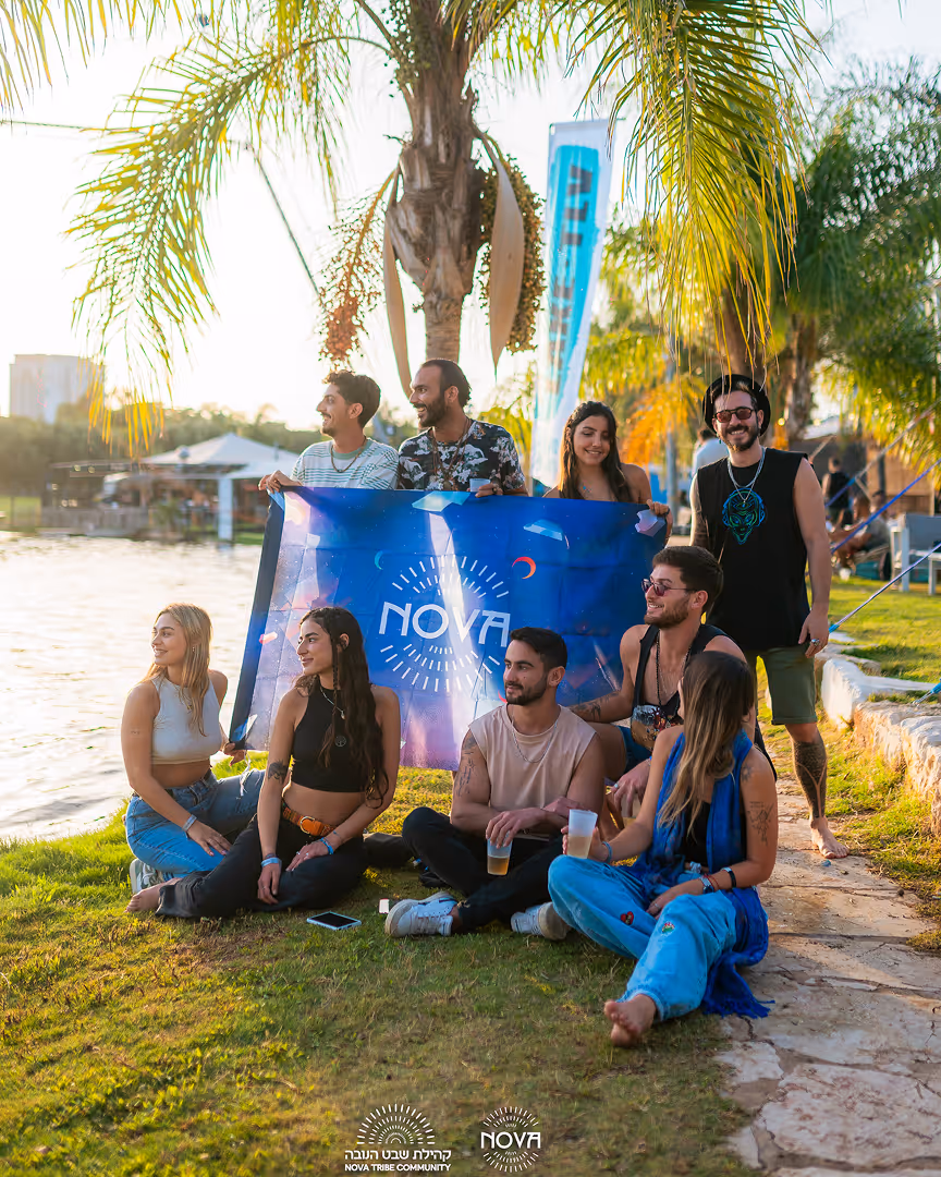 קבוצה של צעירים יושבים ועומדים עם דגל כחול עם סמל NOVA לצד אגם ביום שמש עם עצי דקל ברקע.