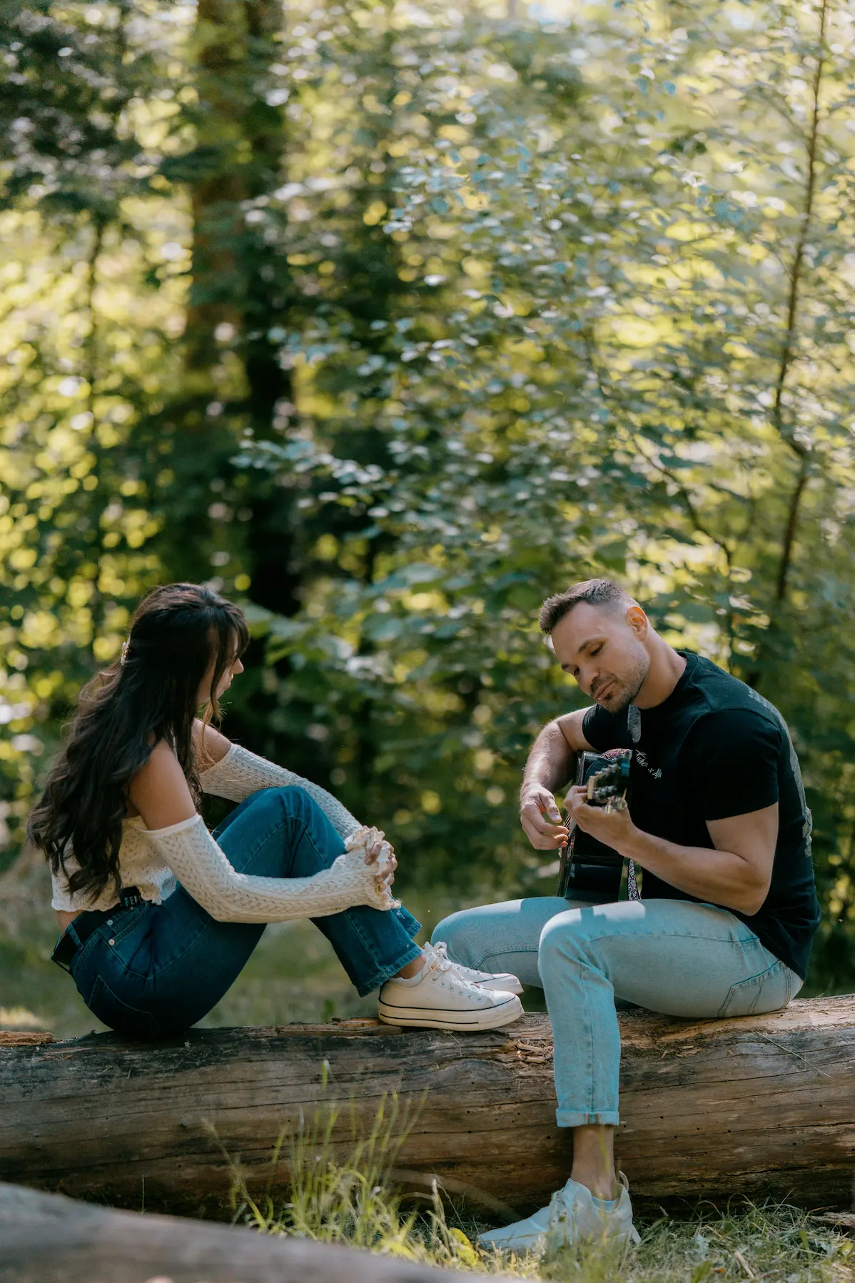 A man plays a guitar while sitting on a fallen log in a forest, facing a woman sitting opposite him. She wears an off-the-shoulder top with long lace sleeves and jeans, looking at him.