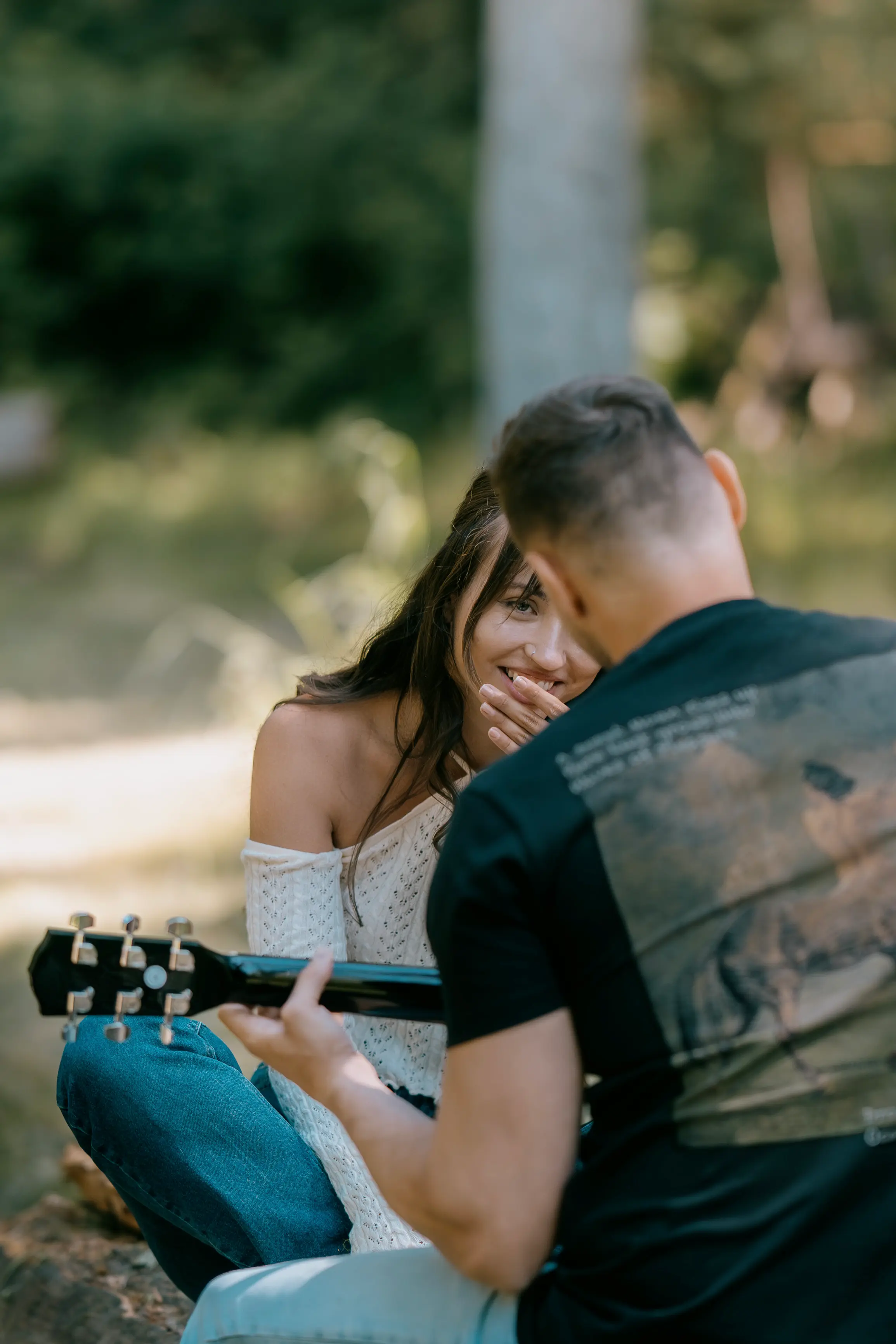 A close-up from behind a man playing a guitar shows a smiling woman with long dark hair covering her mouth with her hands, looking at him. They are sitting outdoors in a softly blurred background.