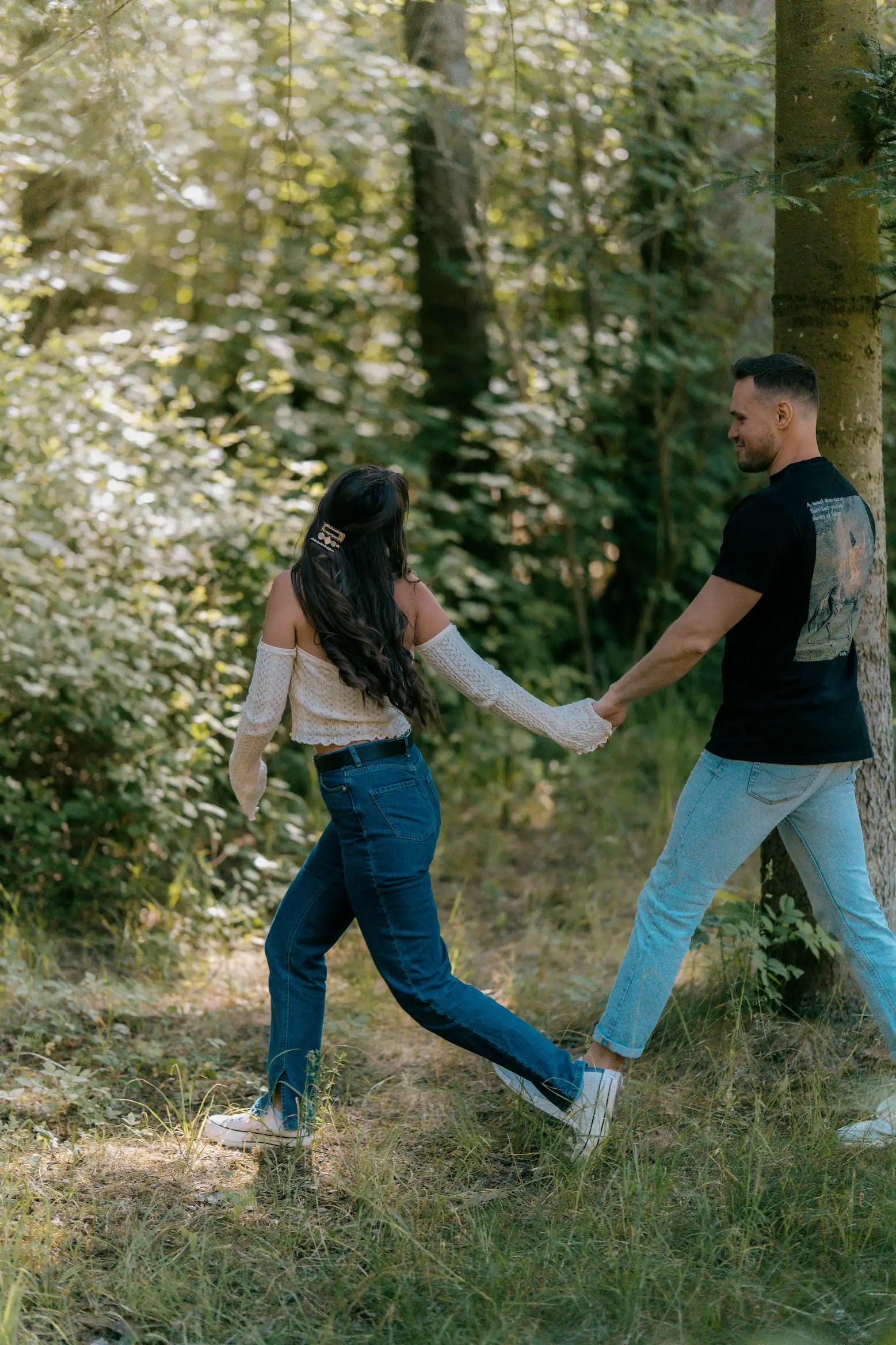 A couple walks hand-in-hand through a grassy forest, away from the viewer. The woman wears an off-the-shoulder top with lace sleeves and jeans, while the man wears a black t-shirt and light jeans.