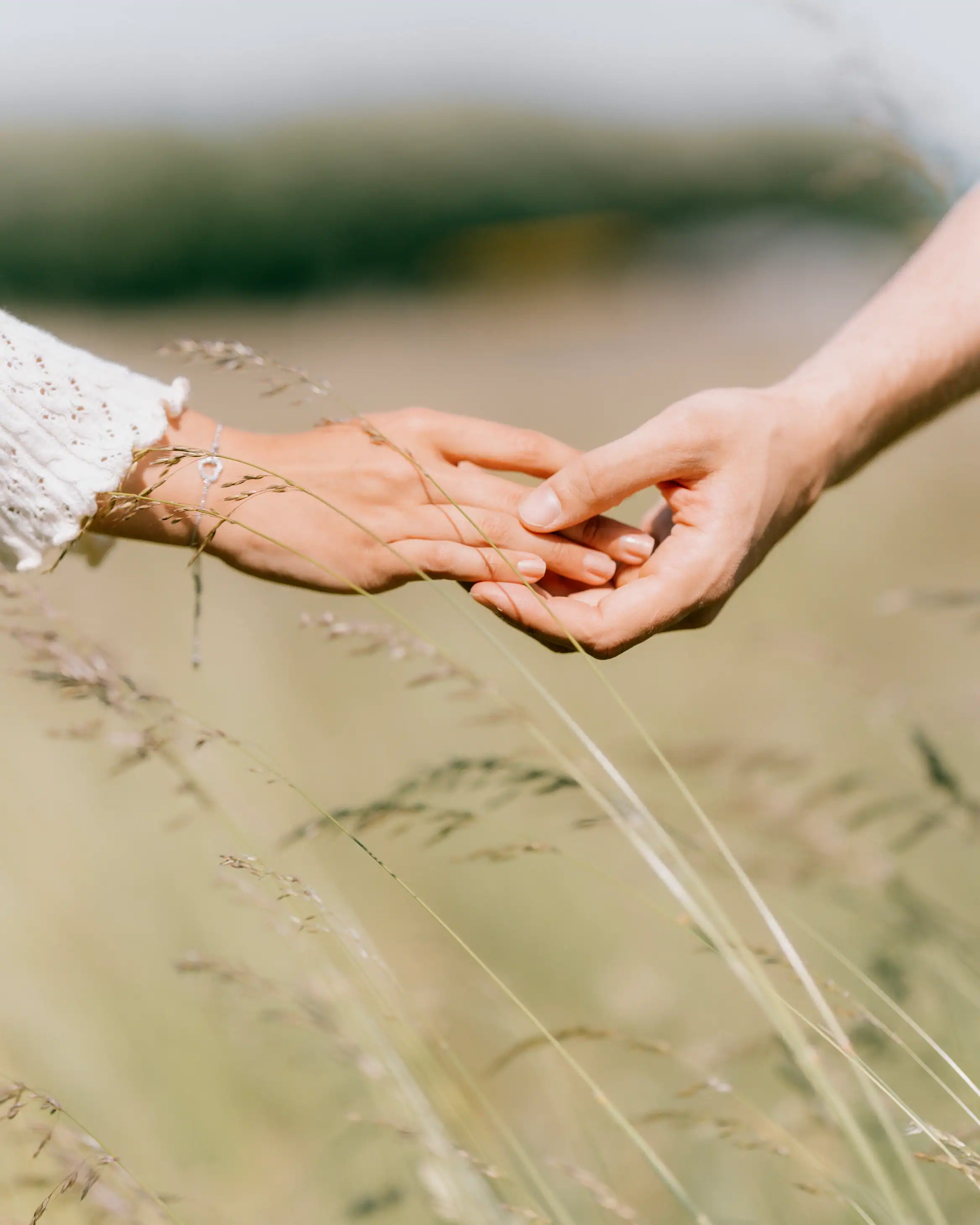 A close-up shows a man and woman gently holding hands amidst tall, dry grass. The woman wears a delicate bracelet and a textured long sleeve.