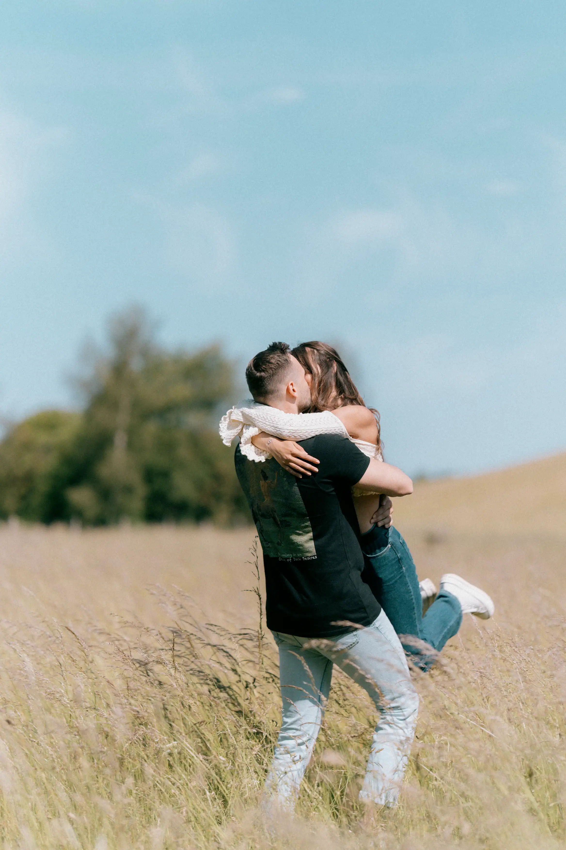 A man lifts and embraces a woman in a grassy field under a bright sky. She wears an off-the-shoulder top with lace sleeves and jeans, wrapping her arms around him.