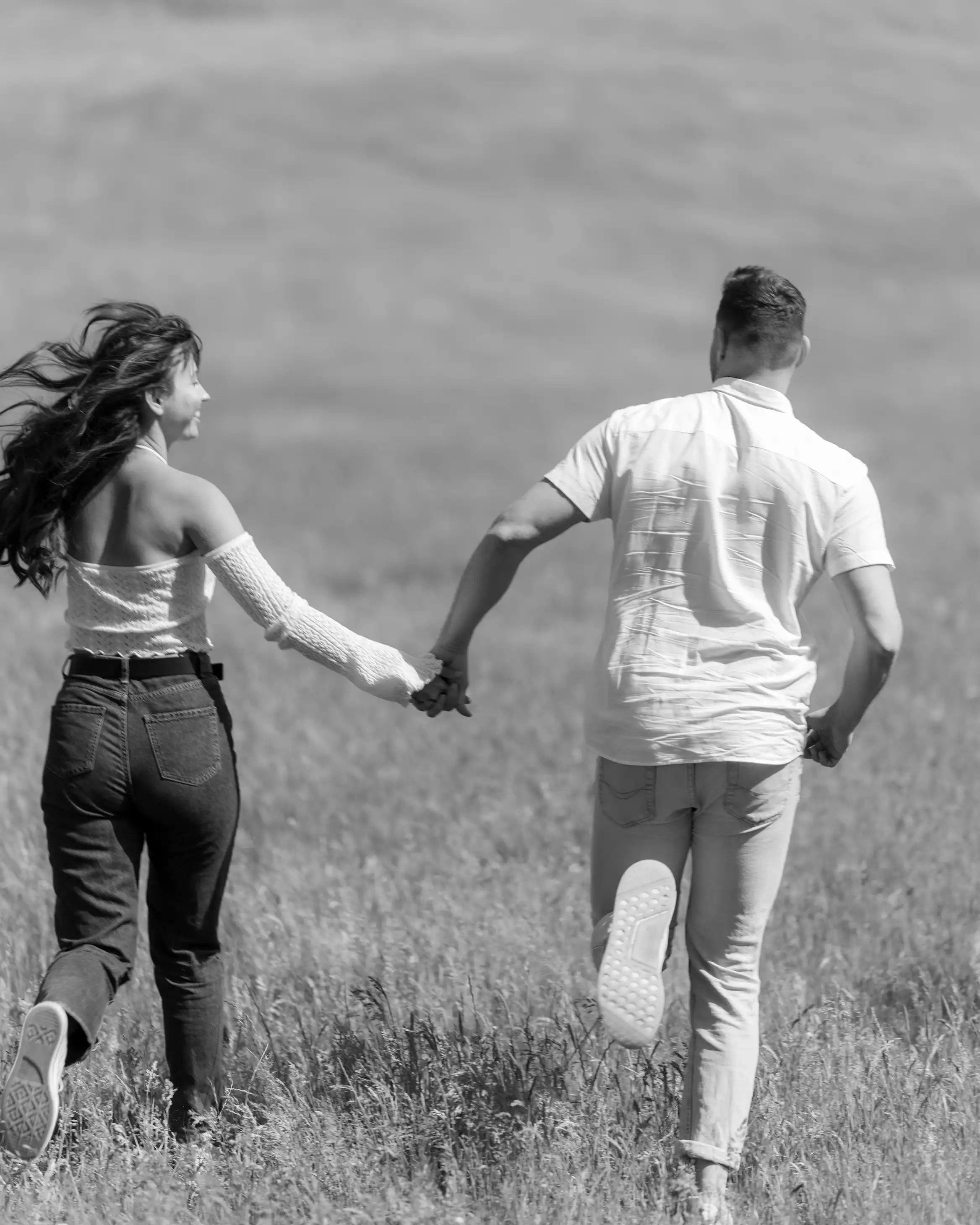 A black and white shot from behind shows a couple running hand-in-hand through a field of tall grass. The woman's long hair flows behind her as she looks back and smiles.