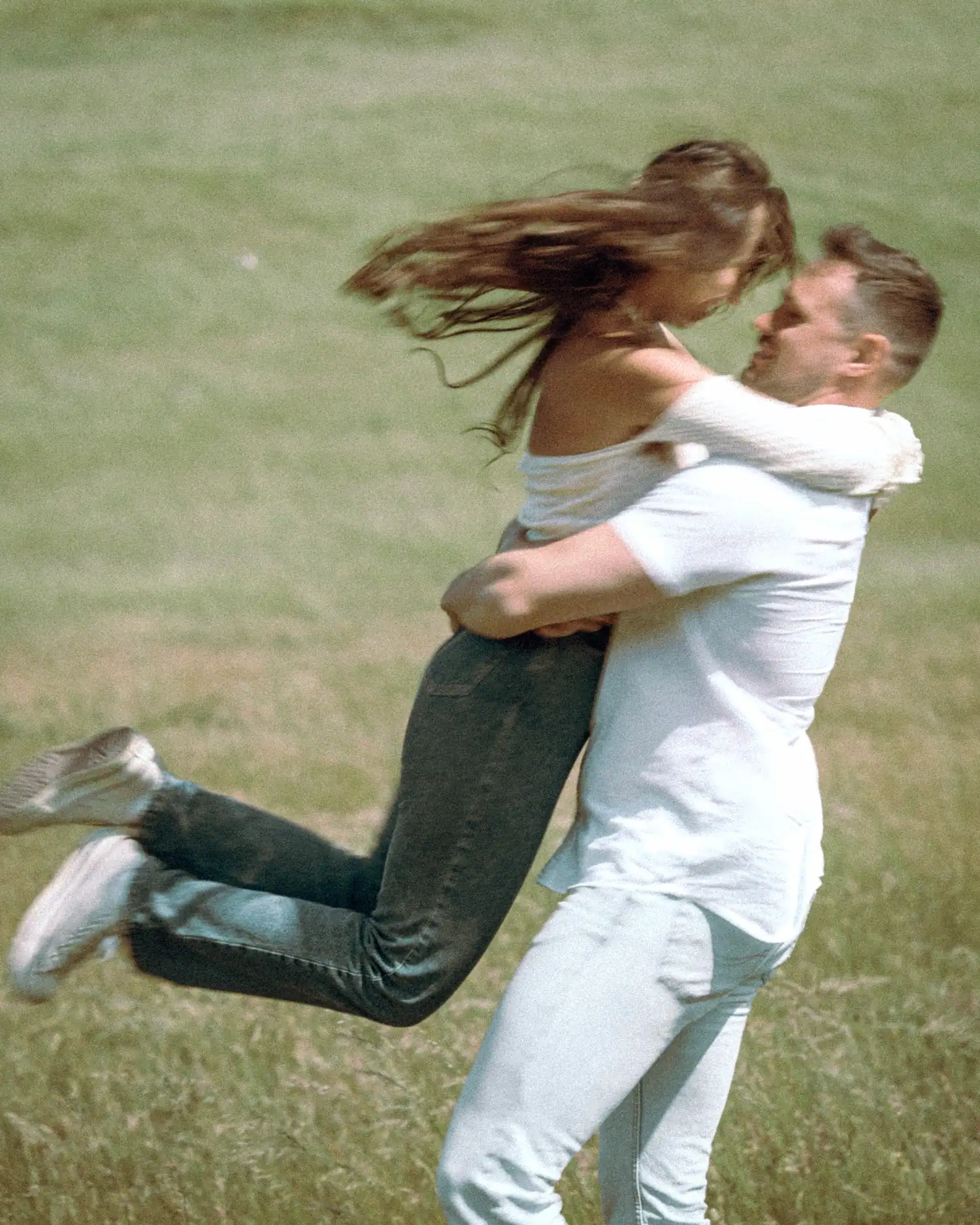 A blurred, vintage-toned image captures a man twirling a woman in his arms in a grassy area. Her hair flies out as she looks at him, wearing an off-the-shoulder top and jeans.