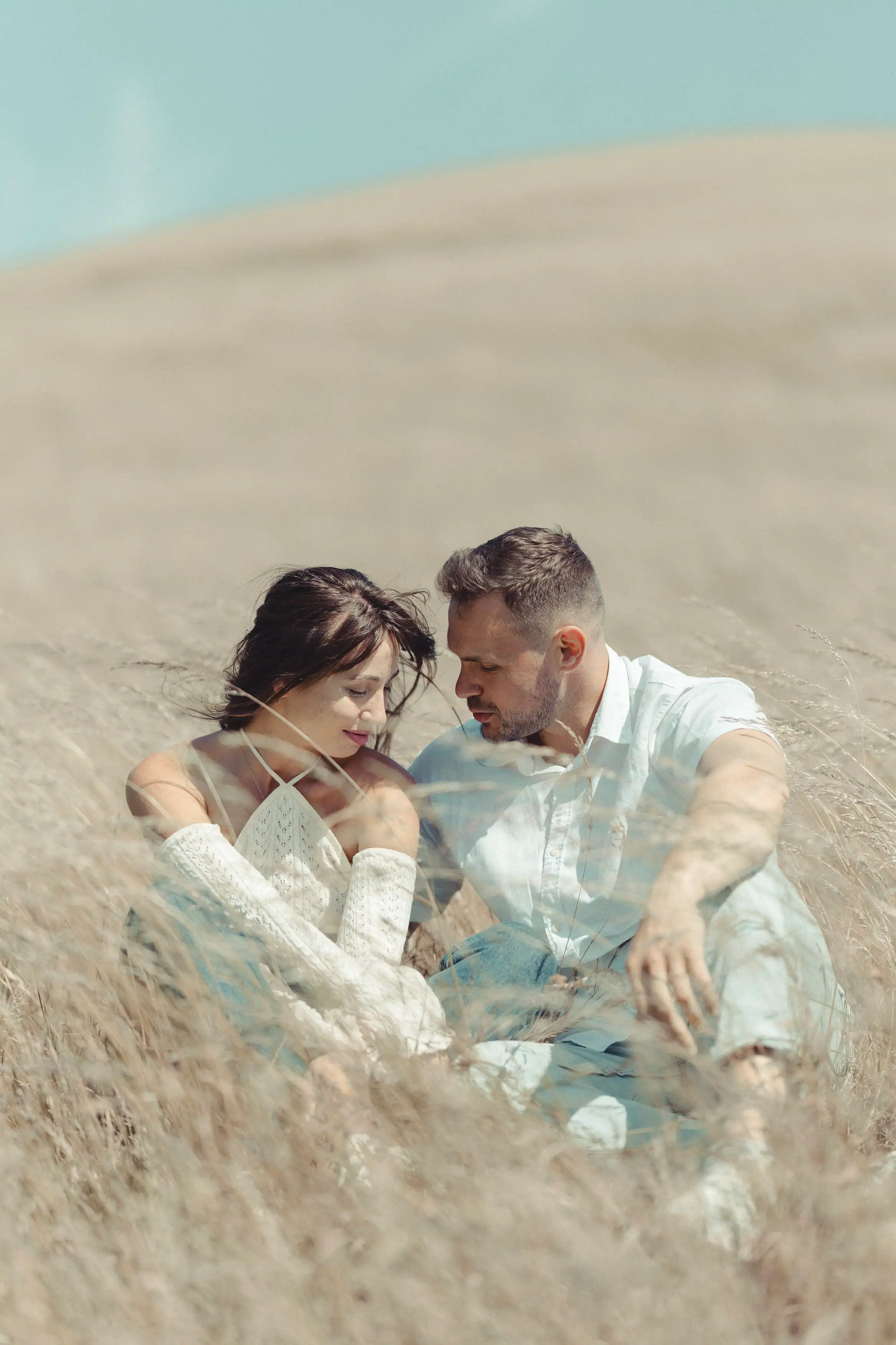 A couple sits close together in a field of tall, dry grass under a clear sky, looking at each other tenderly. The woman wears an off-the-shoulder top with long lace sleeves and jeans.