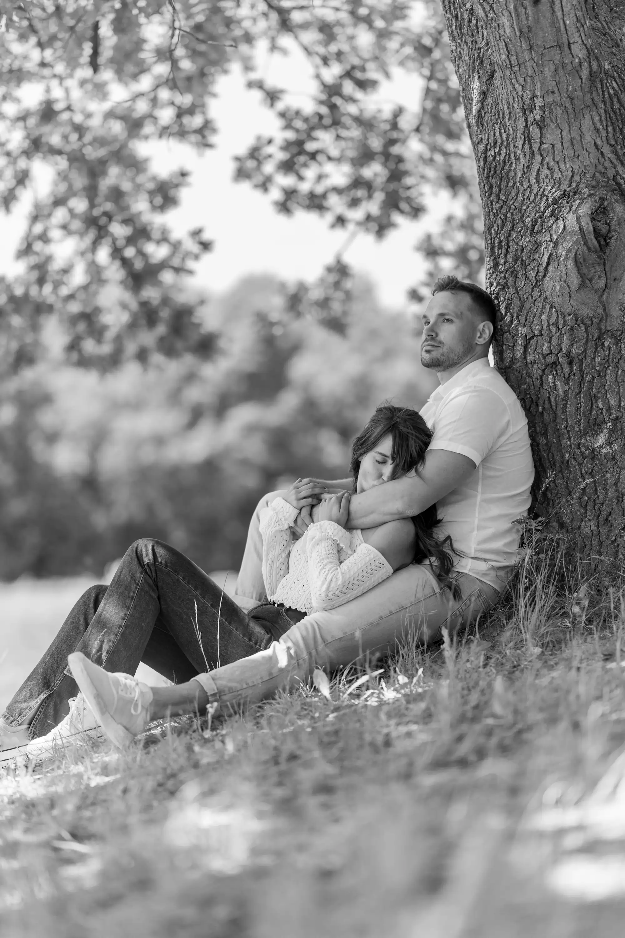 A black and white image shows a man sitting with his back against a large tree, gently embracing a woman who is resting her head on his arm. They are sitting on grassy ground with dappled sunlight filtering through the leaves above.