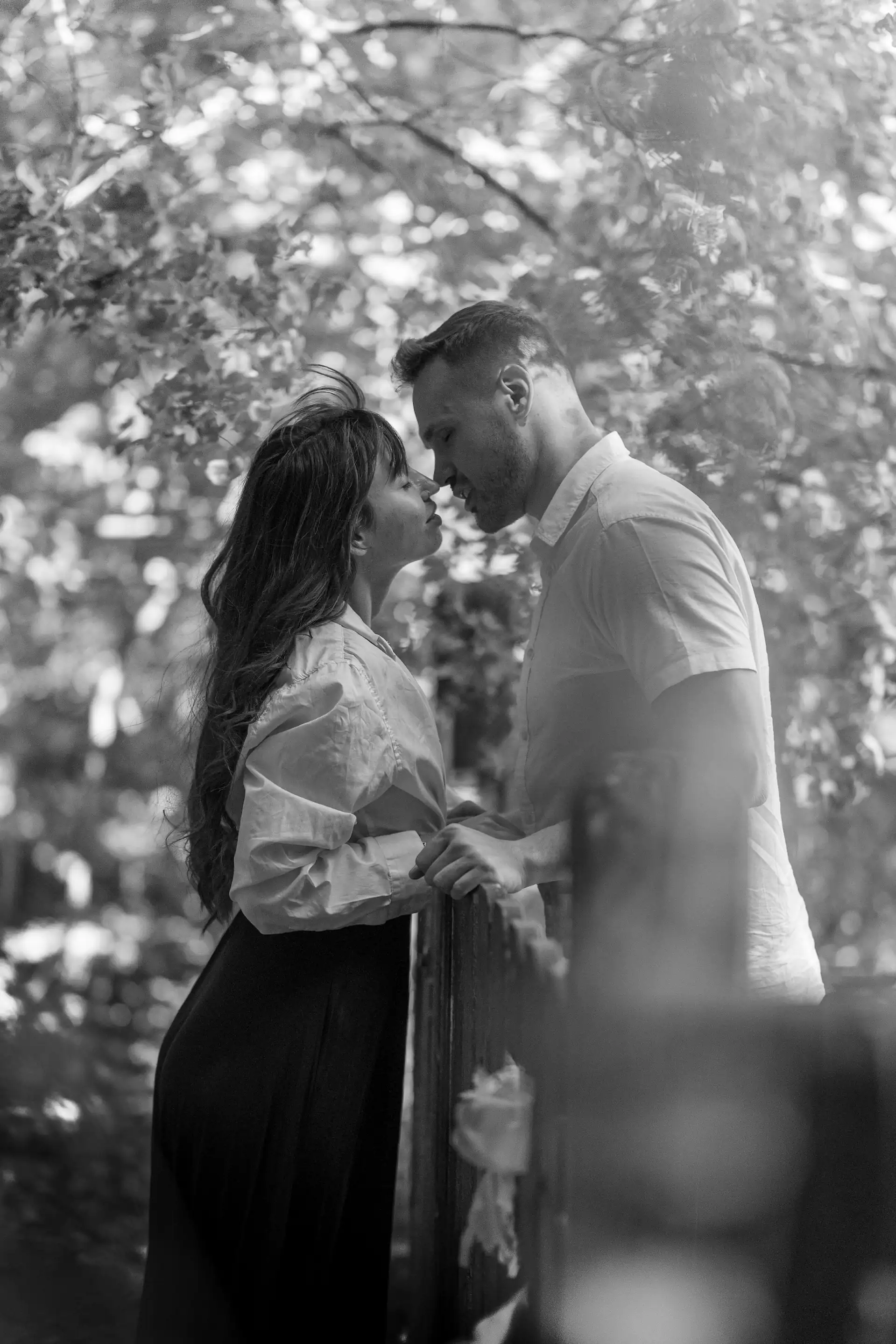 A black and white close-up captures a man and woman standing close to a wooden fence, their faces near each other as if about to kiss. Soft foliage surrounds them, creating a bokeh effect in the background.