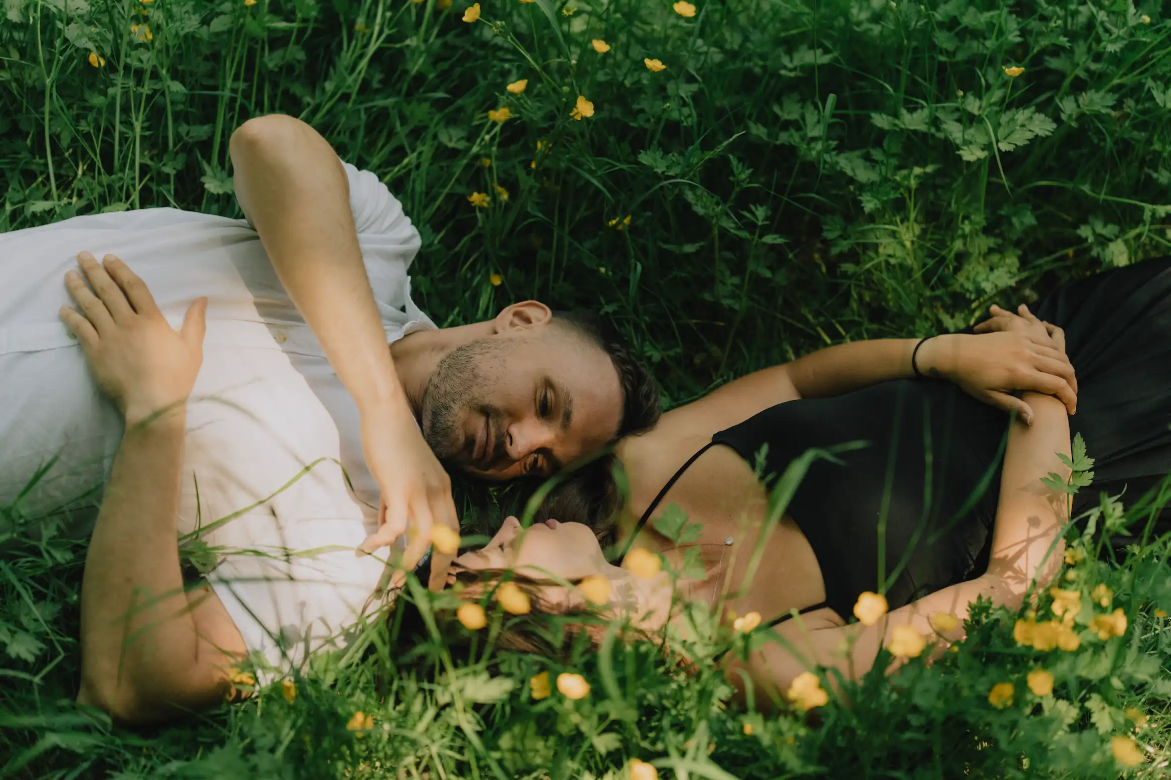 A high-angle shot shows a man and woman lying side-by-side in a field of green grass dotted with small yellow wildflowers. They are looking at each other with relaxed expressions.