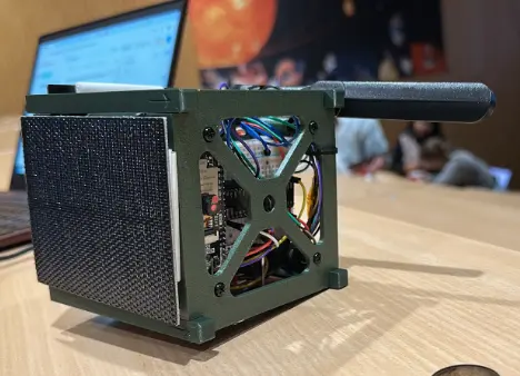 Mini satellite model with visible internal wiring and an extended antenna on a wooden table.