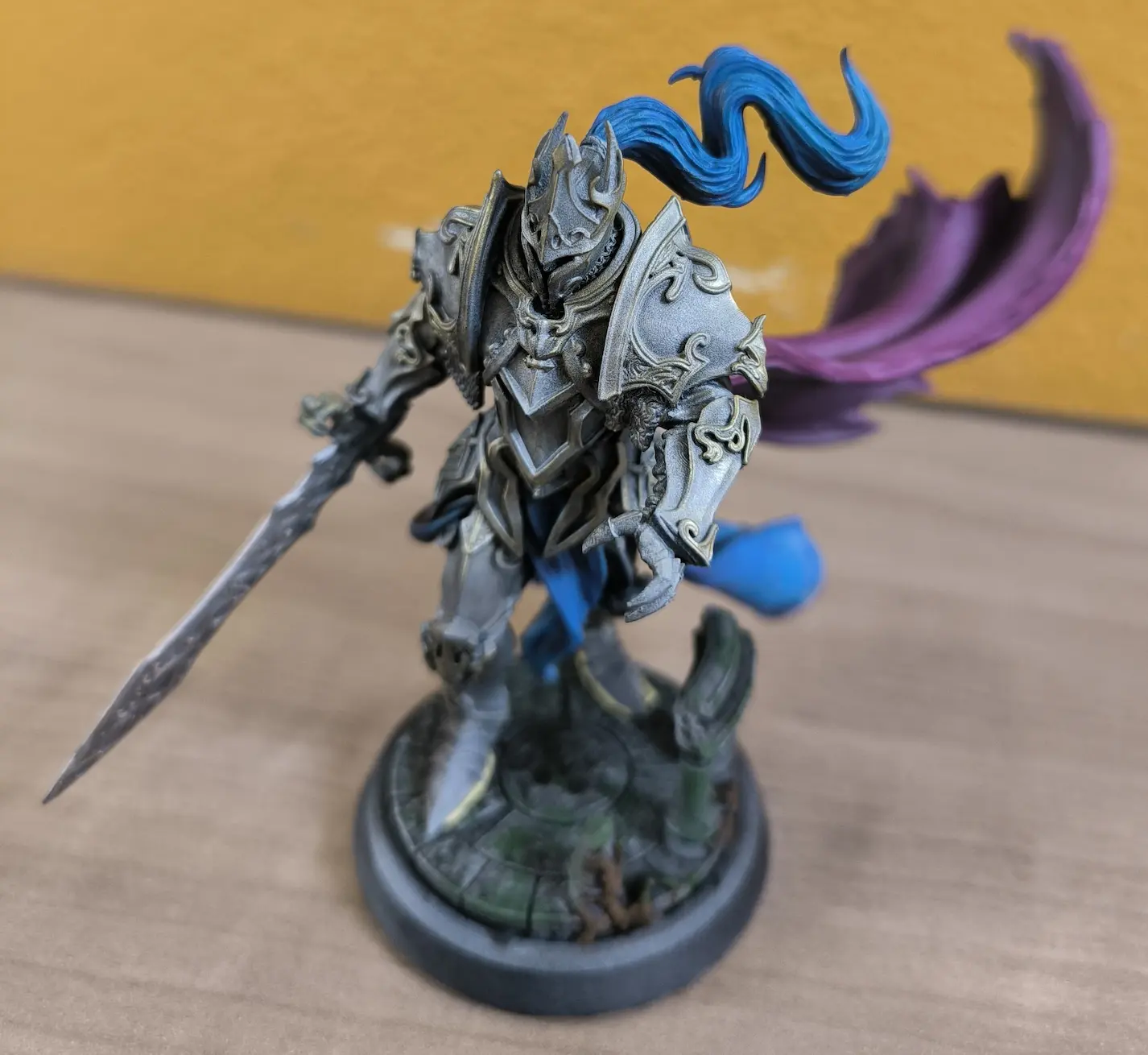 Figurine de chevalier en armure détaillée avec une épée, une cape pourpre et une crinière bleue flottante, sur un socle décoré.