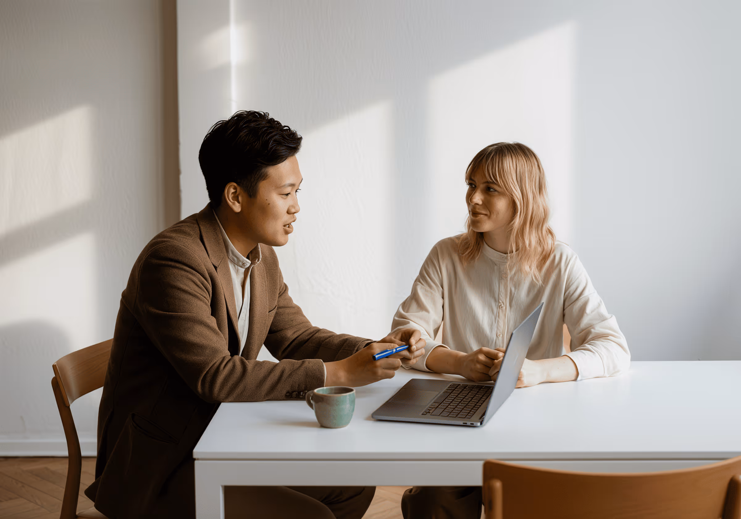 Two people having a discussion at a table with a laptop and cup in a bright room.