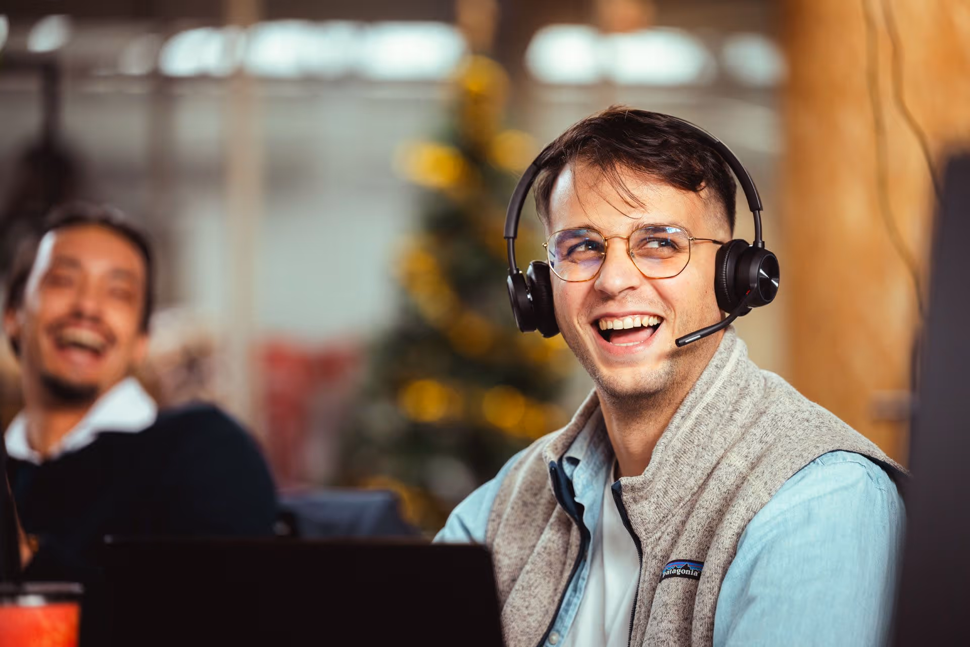 Smiling man with glasses and headset in a casual office setting, another person laughing blurred in background.