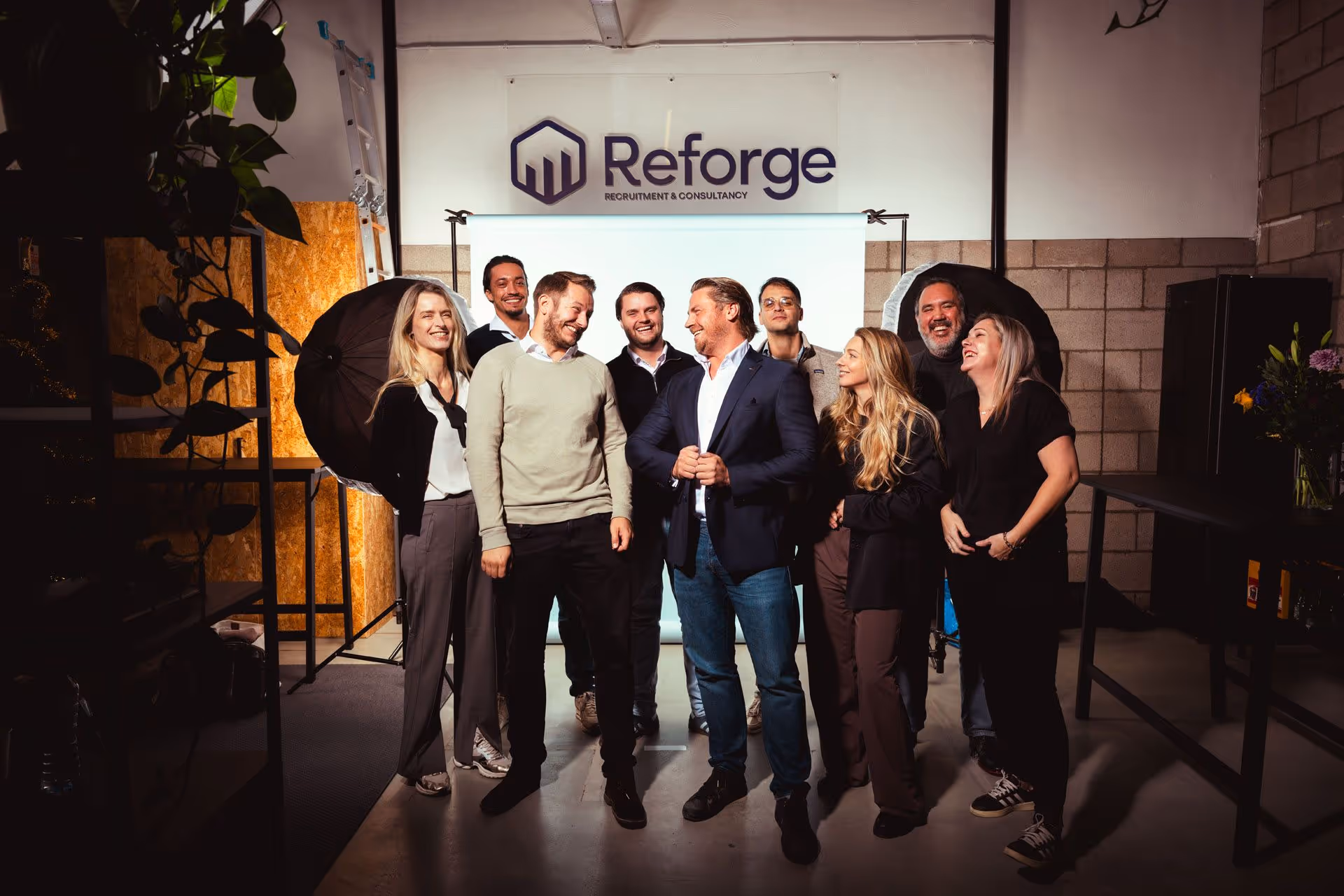 Group of nine diverse people standing, smiling, and interacting in a photo studio with Reforge sign.