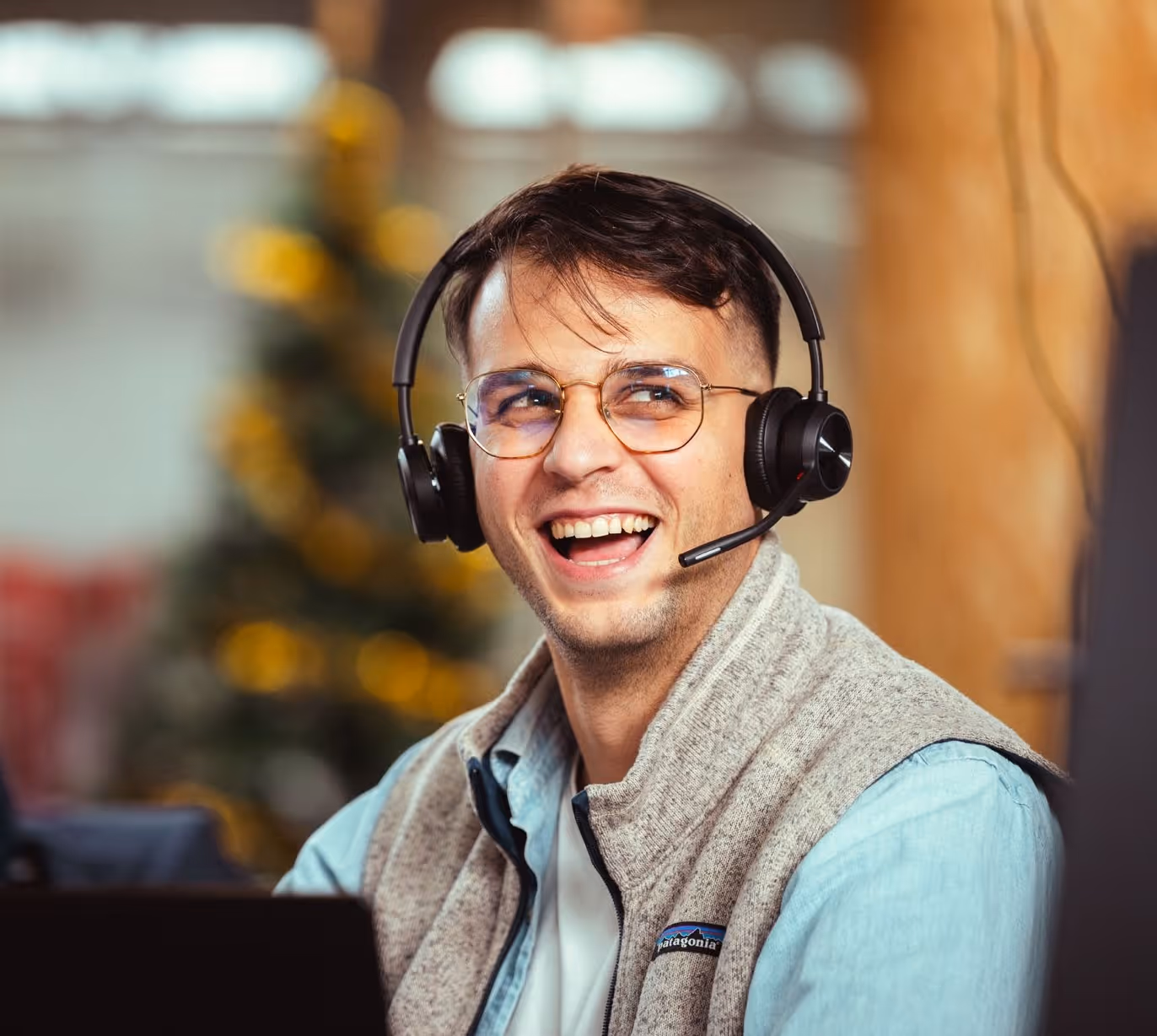 Smiling young man wearing glasses and headset with microphone, working on a laptop indoors.