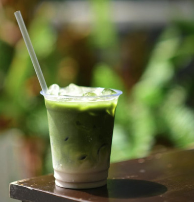 Iced matcha latte layered over milk in a clear cup with a straw, on a wooden table with greenery behind.