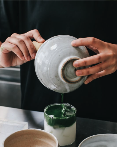 Barista pouring whisked matcha from a spouted bowl over iced milk to build a layered matcha latte.