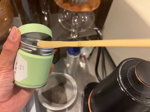 Hand holding an opened matcha tin with a bamboo scoop resting across the rim.