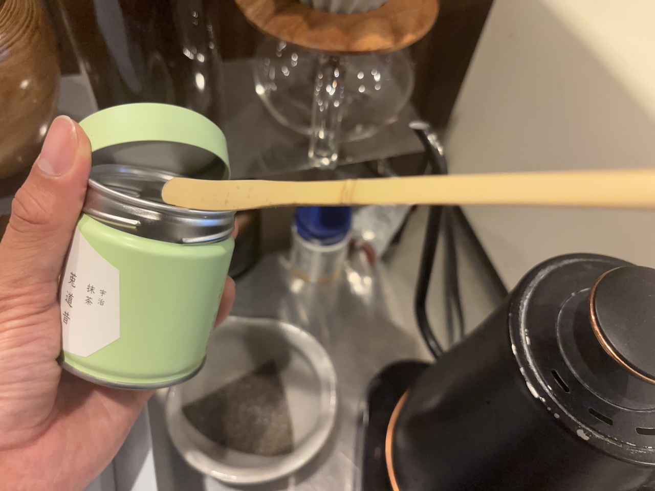 Hand holding an opened matcha tin with a bamboo scoop resting across the rim.