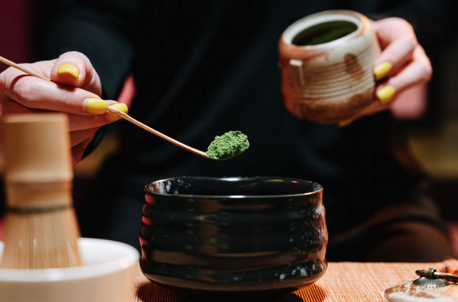 Matcha being measured with a bamboo scoop into a dark tea bowl, with a whisk and caddy alongside.