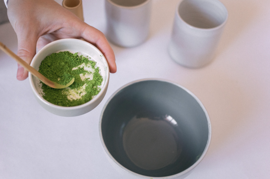 A hand holding a small bowl of bright green matcha powder with a wooden scoop, beside ceramic cups