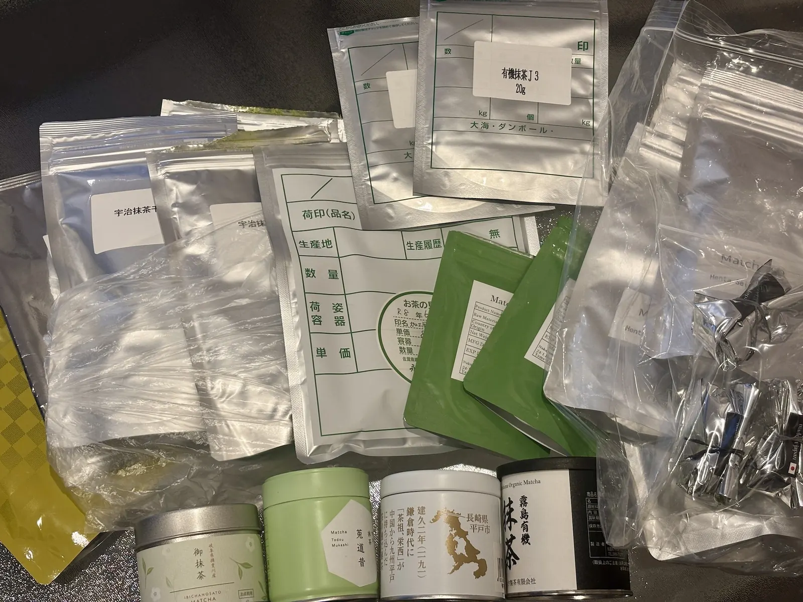 Matcha samples laid out for evaluation: labelled sample bags from Japanese producers, a Japanese product spec sheet, and matcha tins.