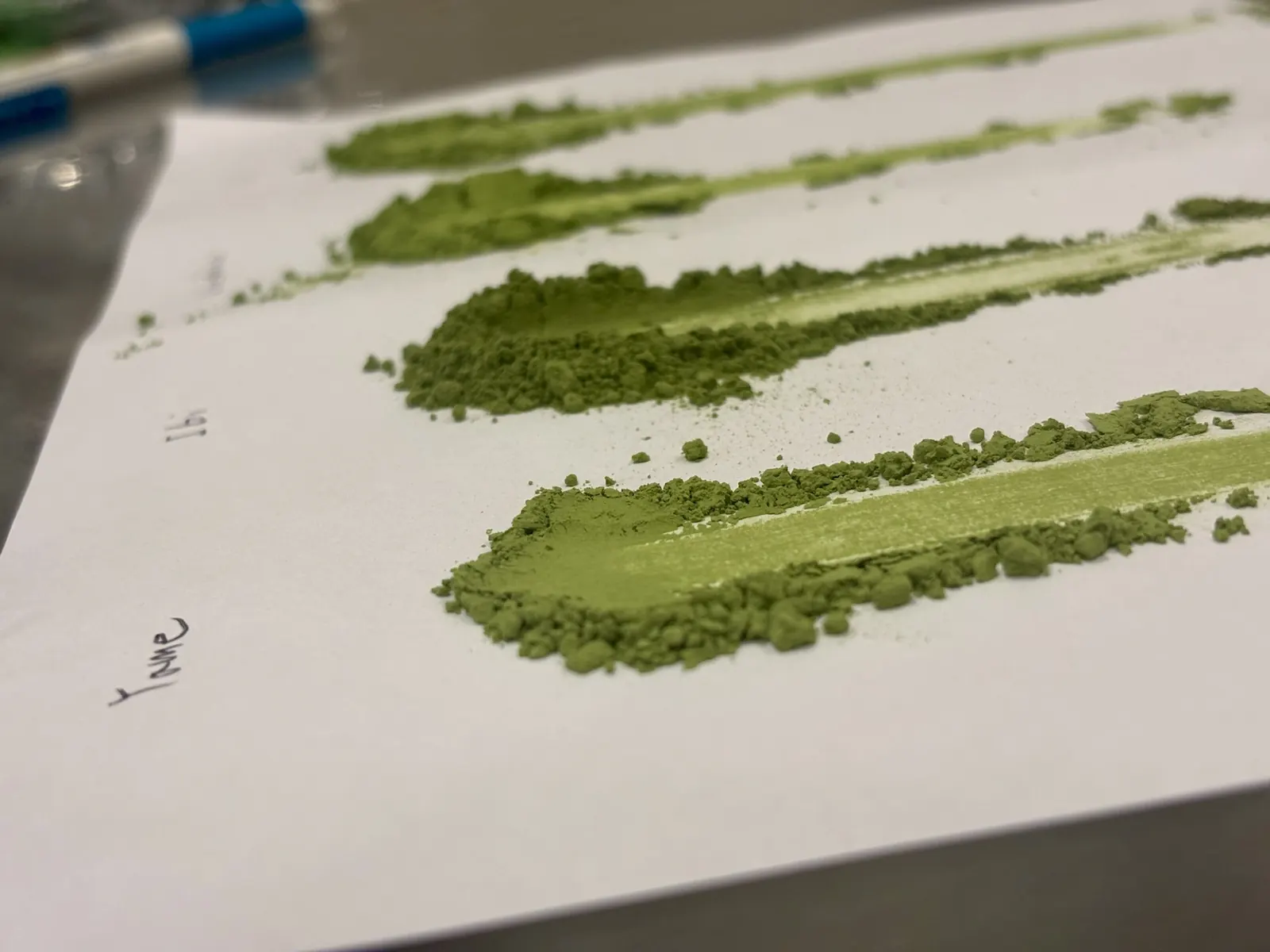 Close-up of matcha samples smeared on paper with handwritten origin labels during a colour comparison.