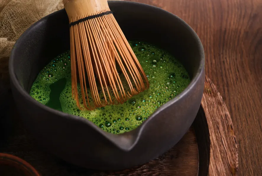 Bamboo whisk resting in a dark spouted bowl of freshly whisked matcha, the concentrate poured over ice.