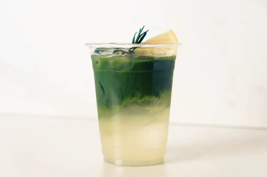Iced matcha lemonade in a clear cup, green matcha layered over cloudy lemon, garnished with rosemary and a lemon wedge.