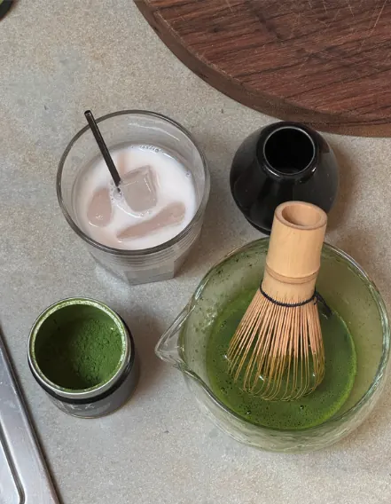 Matcha preparation station: an open caddy of matcha powder, whisked matcha in a spouted glass with a bamboo whisk, and an iced matcha latte.